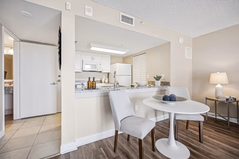 A bright and clean interior space featuring a small dining area with a round white table and two gray upholstered chairs. Behind the dining area is a kitchen with white cabinets, a white refrigerator, a microwave, and a granite countertop with decorative items. To the right, there is a side table with a lamp and some books. The floor is a combination of wood and tile, and a partially open door reveals a bathroom with a granite countertop.