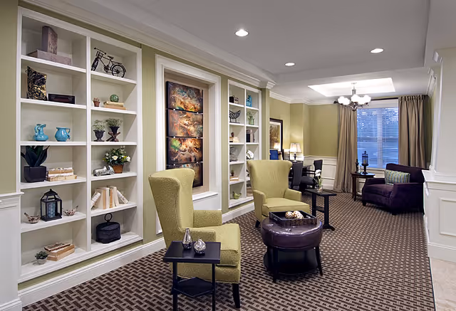 A well-lit senior living lounge with built-in white shelving, two green armchairs, a round ottoman and additional seating by a window.