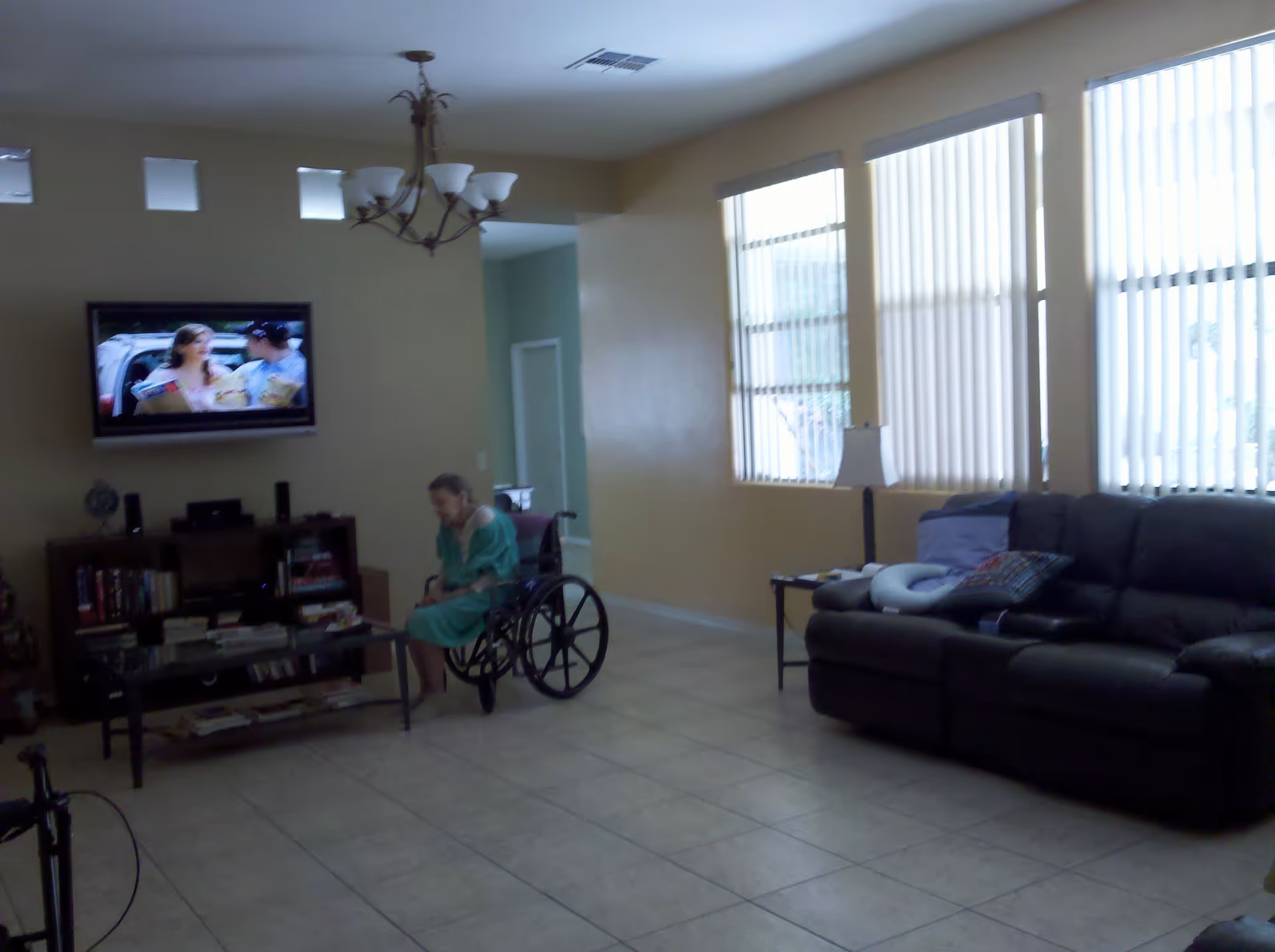 A living room with tiled floor, a black leather couch with pillows, a side table with a lamp, a coffee table, and a bookshelf with books and other items. A person in a green hospital gown is sitting in a wheelchair near the coffee table. A flat-screen TV is mounted on the wall showing a scene with two people. Large windows with vertical blinds allow natural light into the room.
