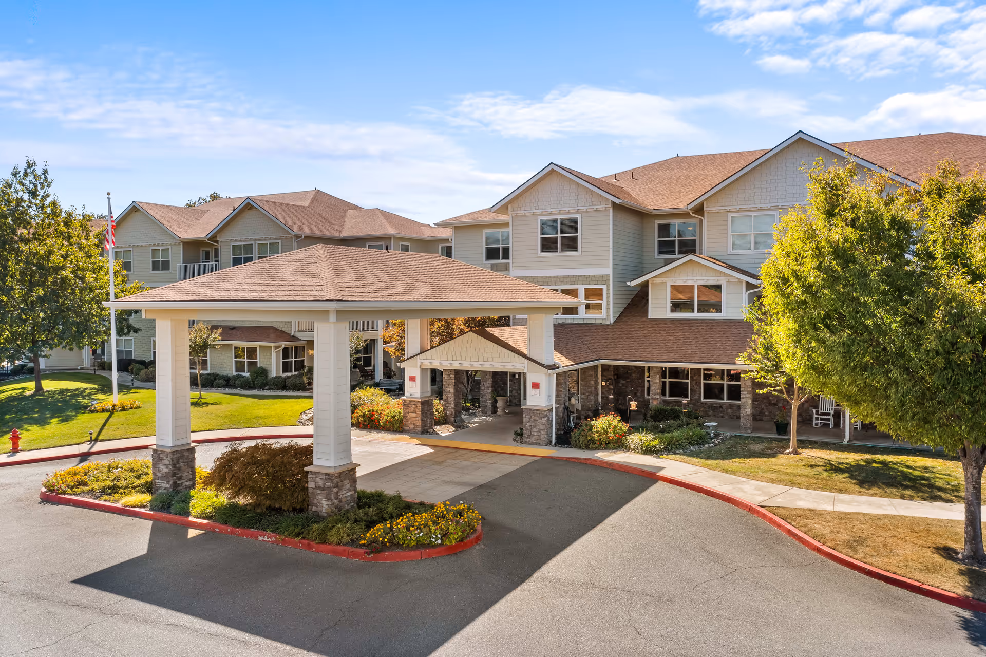 Front exterior view of Shasta Estates Senior Living facility showing a large covered entrance with stone pillars, surrounded by well-maintained landscaping, trees, and a clear blue sky.