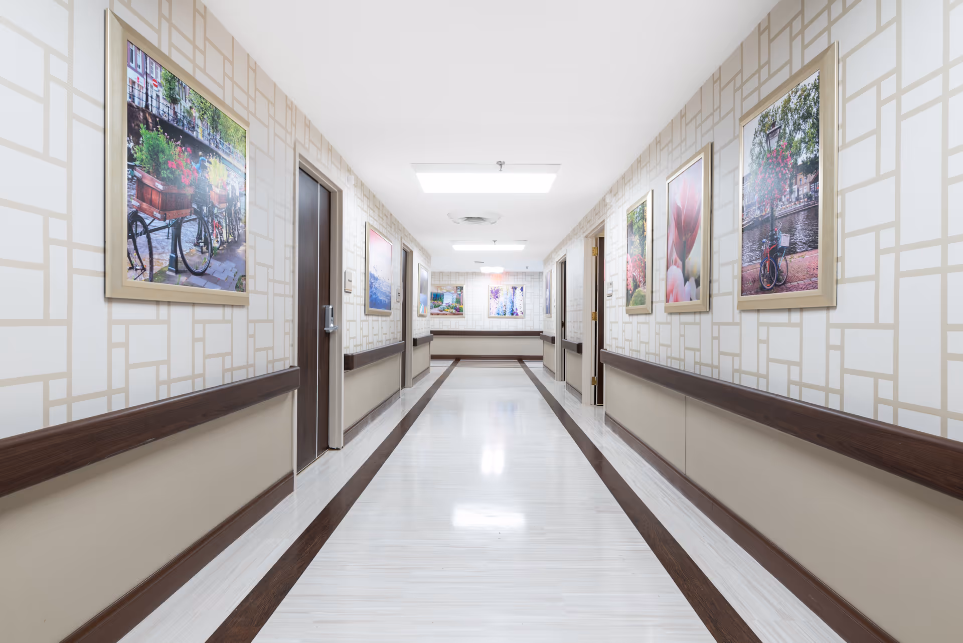 A clean, well-lit hallway in a senior living facility with patterned wallpaper, handrails on both sides, several closed doors, and framed pictures of bicycles and flowers hanging on the walls.