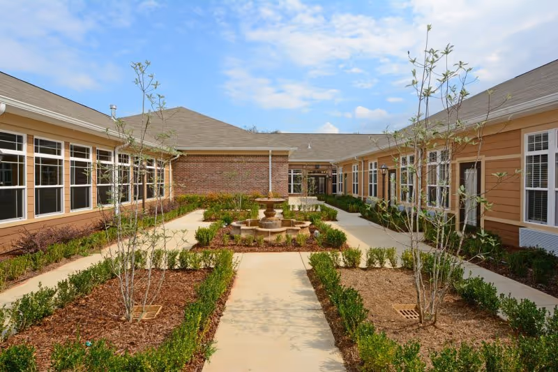 Sunlit landscaped courtyard with a central fountain, walkways, young trees and low hedges between single-story brick and siding buildings.