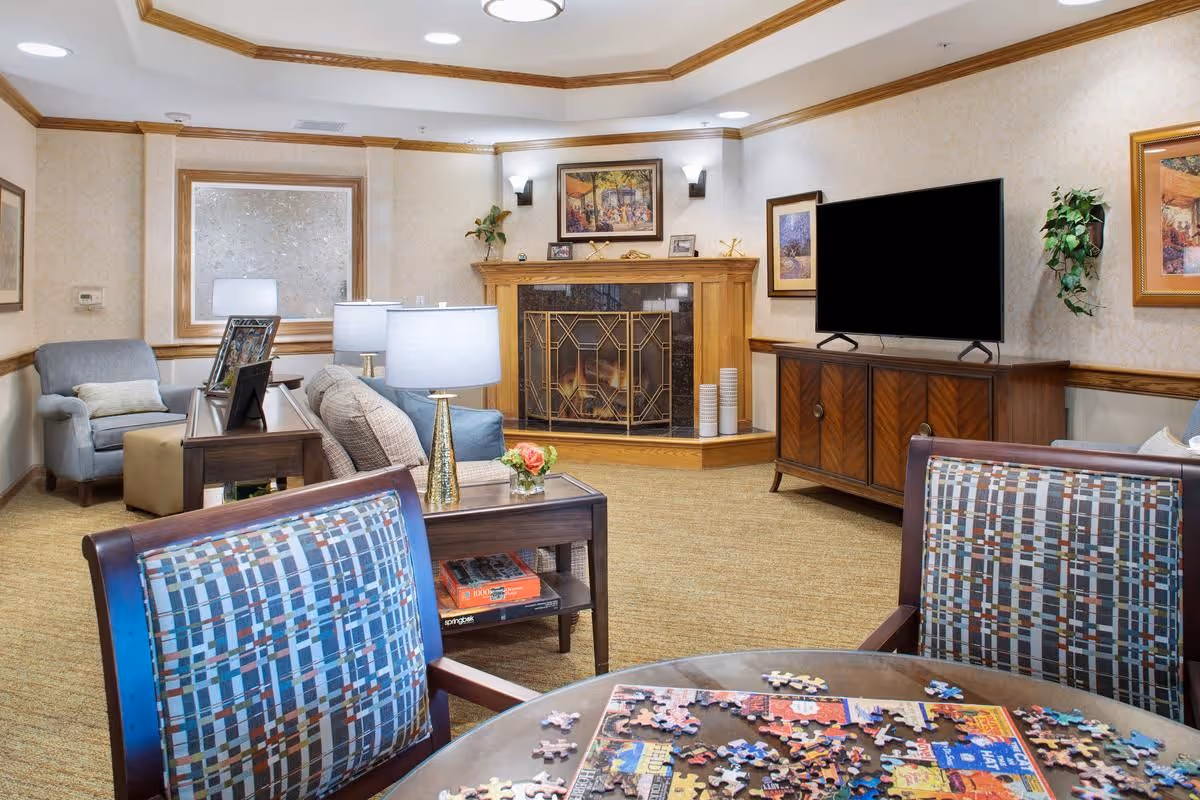 A cozy living room area with a fireplace in the corner, a flat-screen TV on a wooden cabinet, and comfortable seating including armchairs and a sofa. A round table in the foreground has a partially completed jigsaw puzzle on it. The room is decorated with framed artwork, plants, and table lamps, creating a warm and inviting atmosphere.