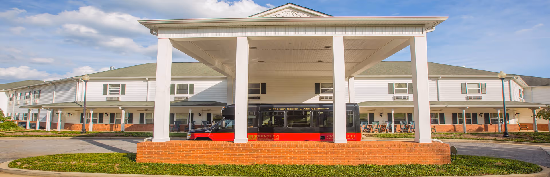 Front entrance of a senior living building with a covered porte-cochere and a red-and-black shuttle bus parked beneath it.