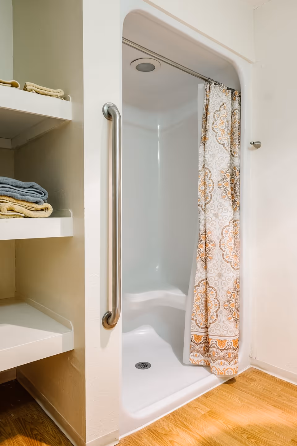 A small shower stall with a white base and a patterned shower curtain in shades of orange and beige. There is a metal grab bar mounted on the wall next to the shower entrance. To the left, there are built-in shelves holding folded towels in yellow and blue. The floor outside the shower is wooden.
