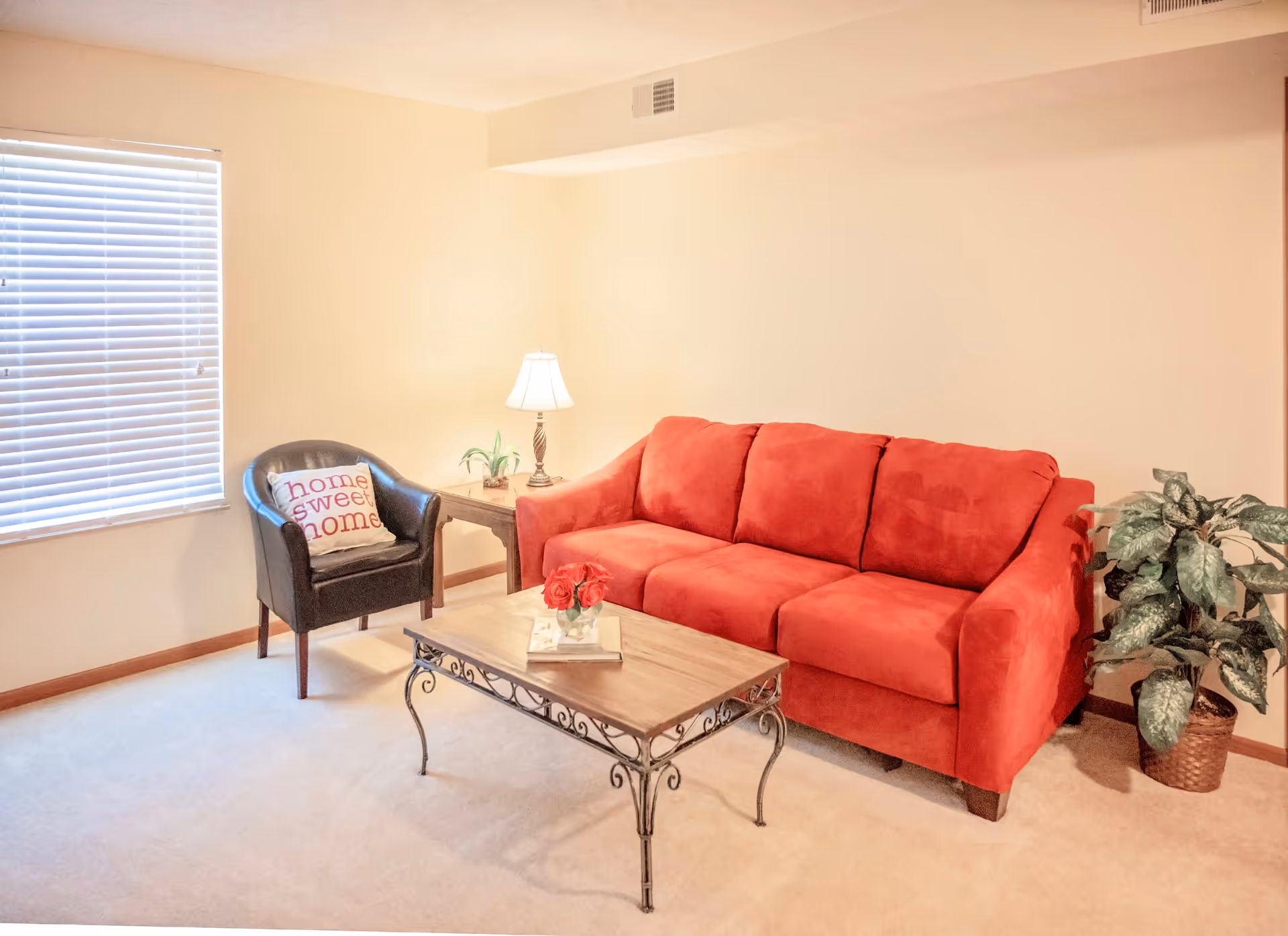 A cozy living room with a bright red sofa, a black armchair with a 'home sweet home' pillow, a wooden coffee table with a small flower arrangement and book, a side table with a lamp and small plant, and a potted plant in the corner. The room has beige walls and carpet, and a window with closed blinds.