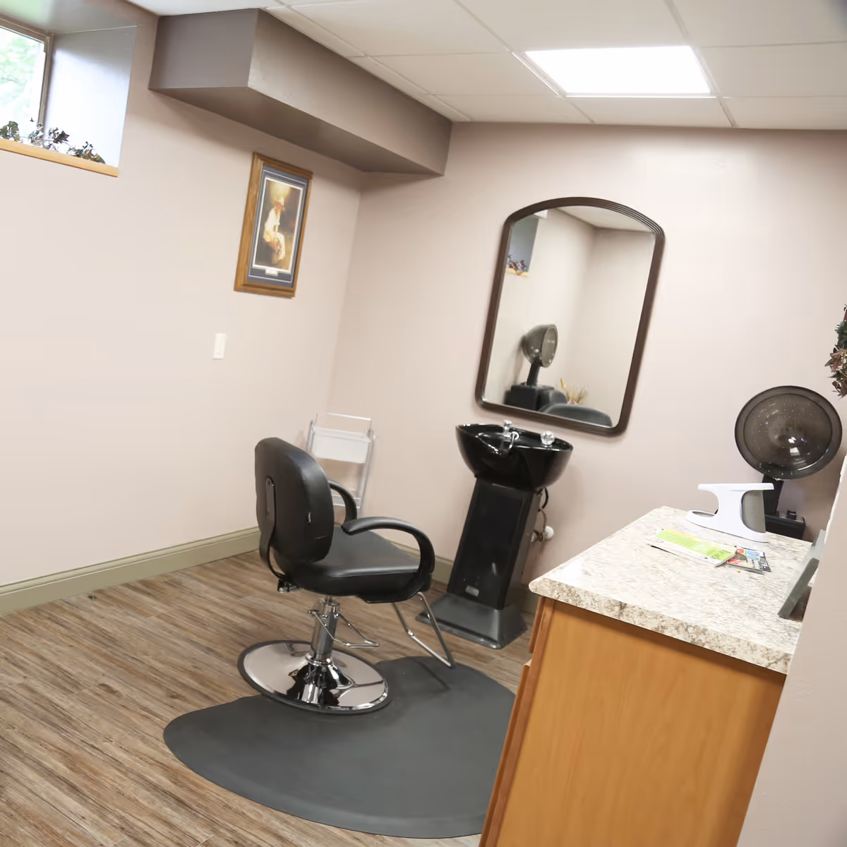 A small hair salon area inside a facility with a black salon chair on a protective mat, a black hair washing sink, a large wall mirror, a countertop with some items on it, and a hair dryer hood in the corner. The room has light-colored walls, wood flooring, and a small window near the ceiling.