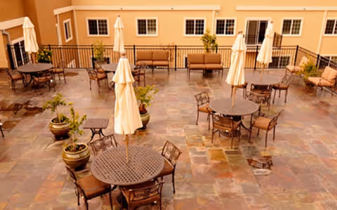Outdoor courtyard patio with round tables, chairs, umbrellas, and potted plants in front of a multi-story building.