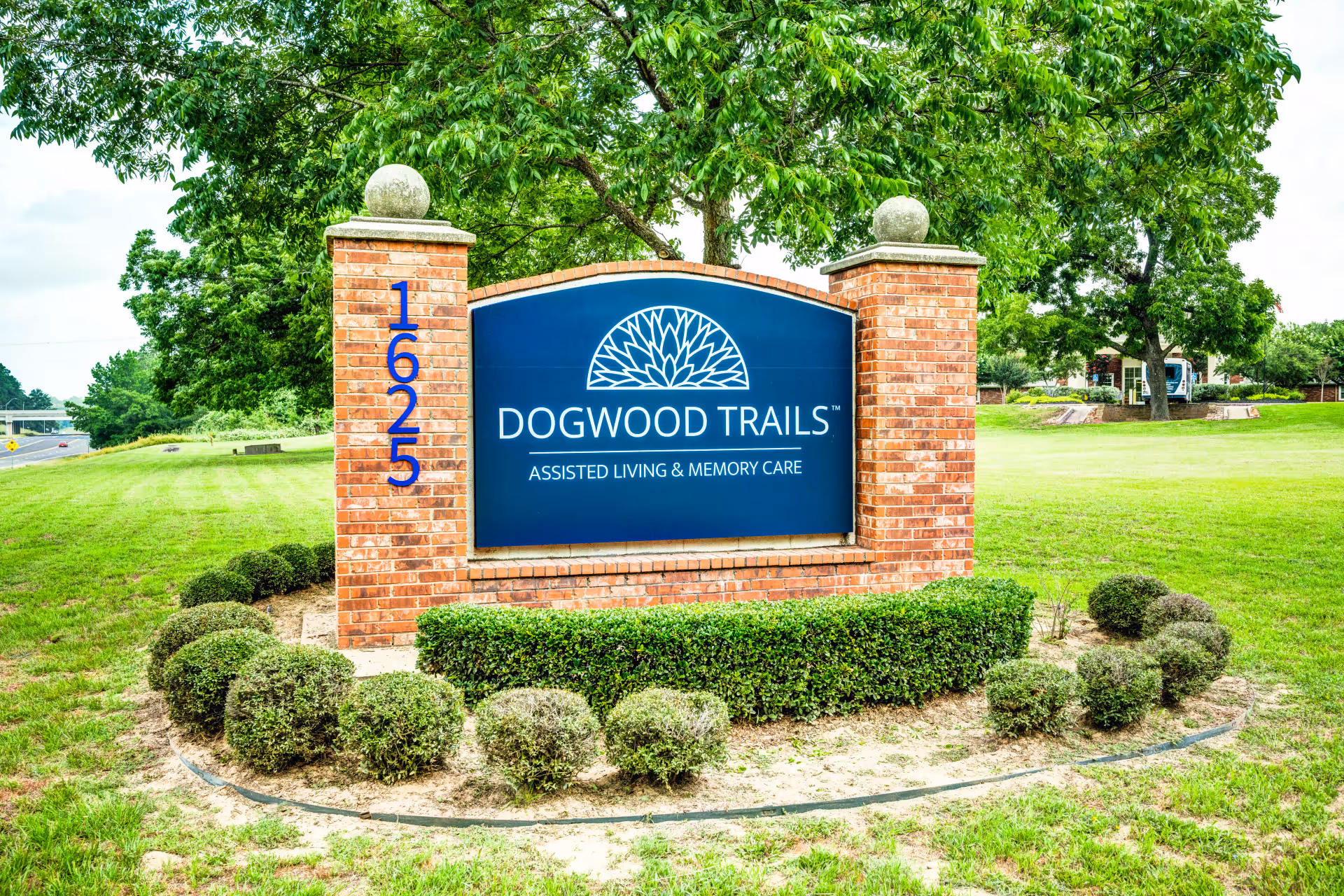 A brick signpost with a blue sign that reads 'DOGWOOD TRAILS ASSISTED LIVING & MEMORY CARE' surrounded by neatly trimmed bushes and green grass with trees in the background.