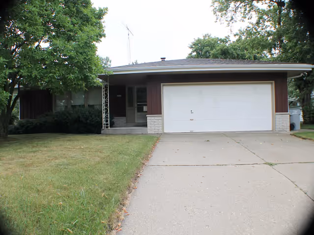 Front exterior view of a single-story house with a two-car garage, a concrete driveway, and a small front porch. The house has brown siding with white brick accents and is surrounded by green grass and trees.