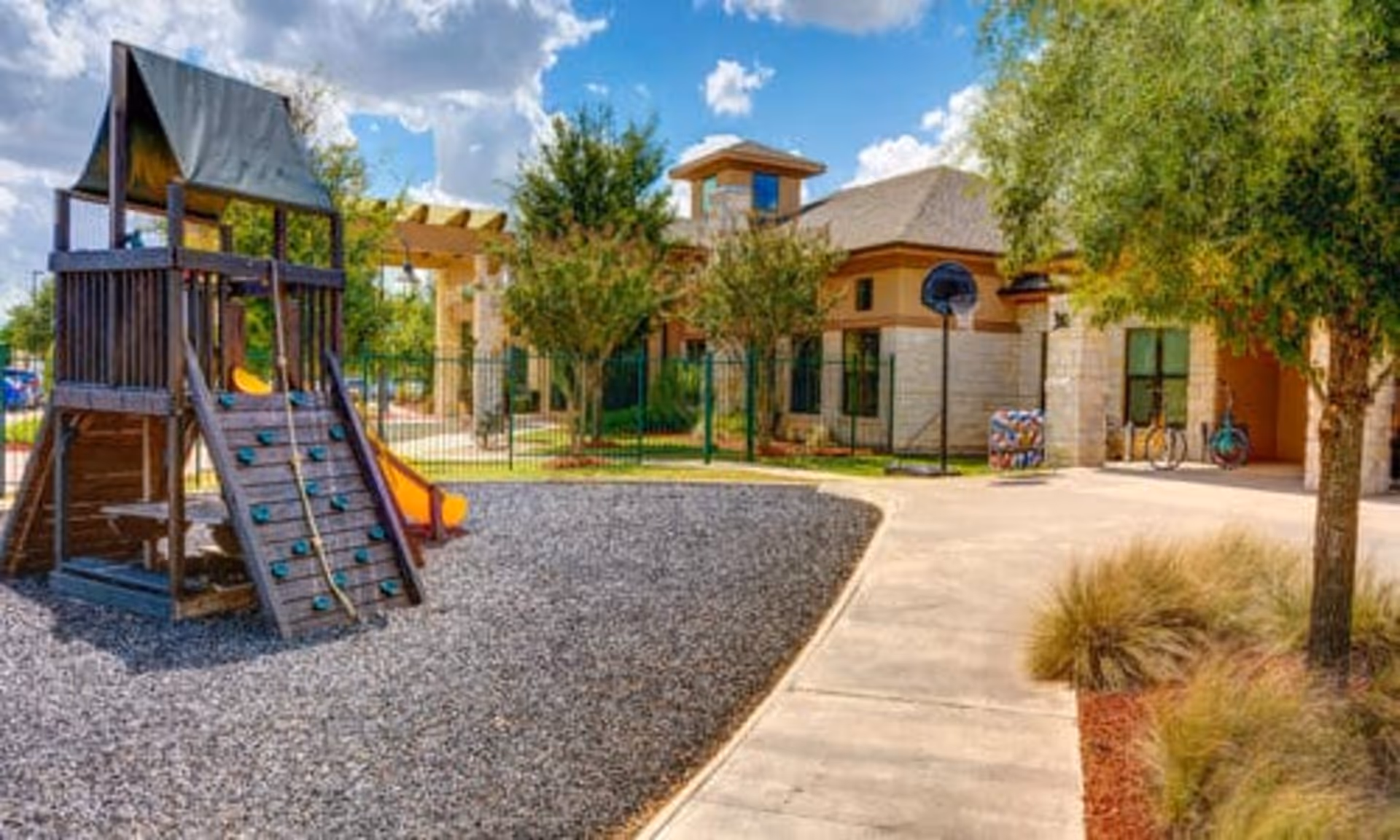 Outdoor playground with a wooden playset and slide in front of a one-story building and walkway under a partly cloudy sky.