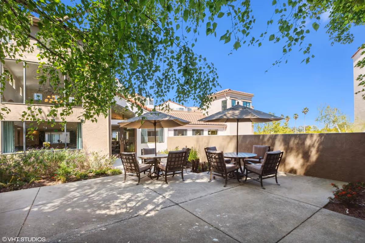 Outdoor patio area at Cadence Millbrae with two round tables, each surrounded by four cushioned chairs and shaded by large umbrellas. The patio is paved with concrete and bordered by a low wall, with greenery and trees providing shade and a view of the building's exterior under a clear blue sky.