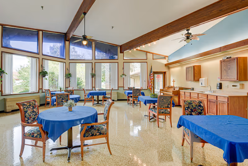 A bright dining room with large windows letting in natural light. The room has several round tables covered with blue tablecloths and floral upholstered chairs. There are hanging plants by the windows, wooden cabinets, a piano, and an American flag in the corner.