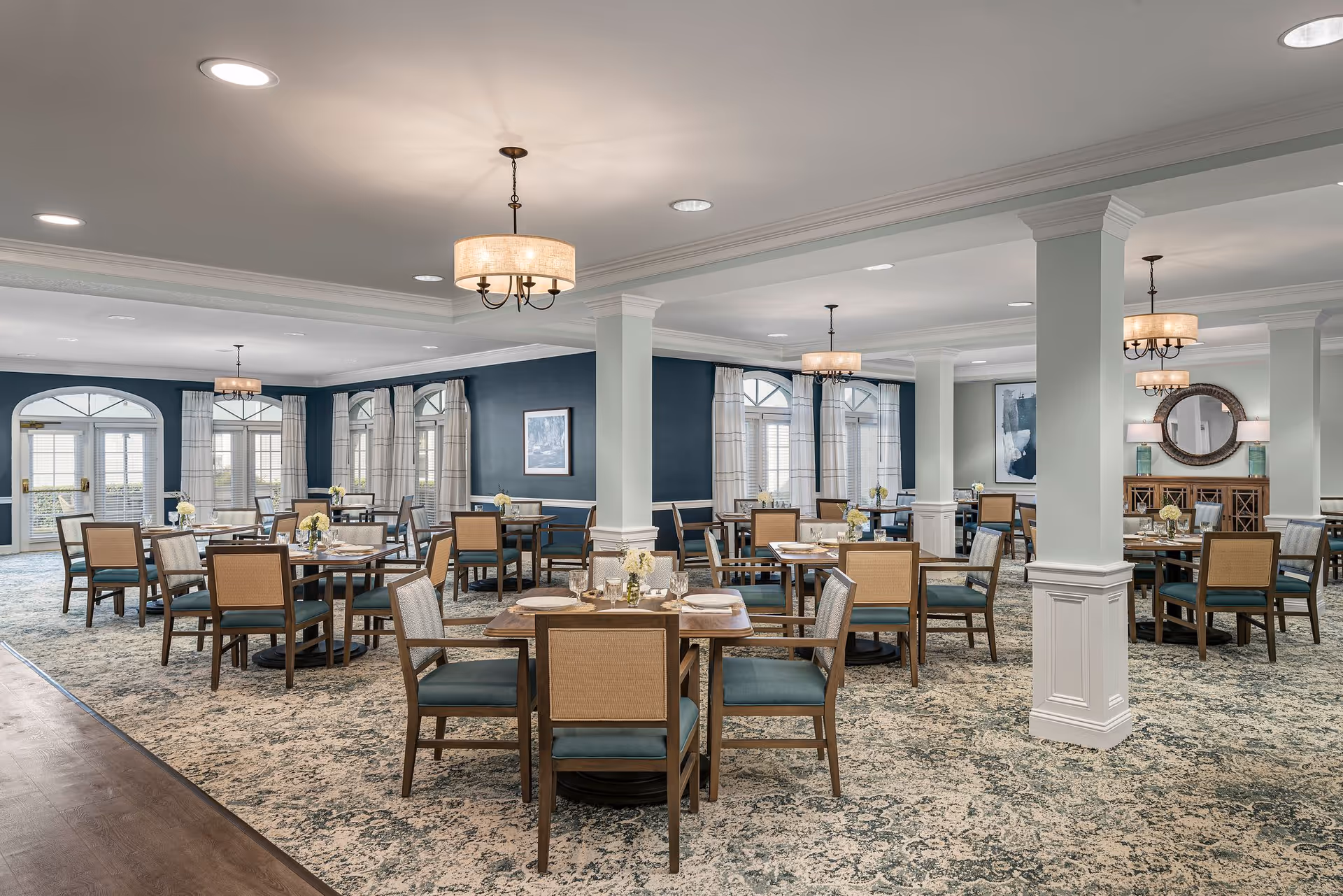A spacious dining room with multiple wooden tables and chairs arranged neatly. The room features large windows with white curtains, blue accent walls, patterned carpet, and elegant ceiling lights. There are decorative elements including framed artwork and a round mirror on the walls.