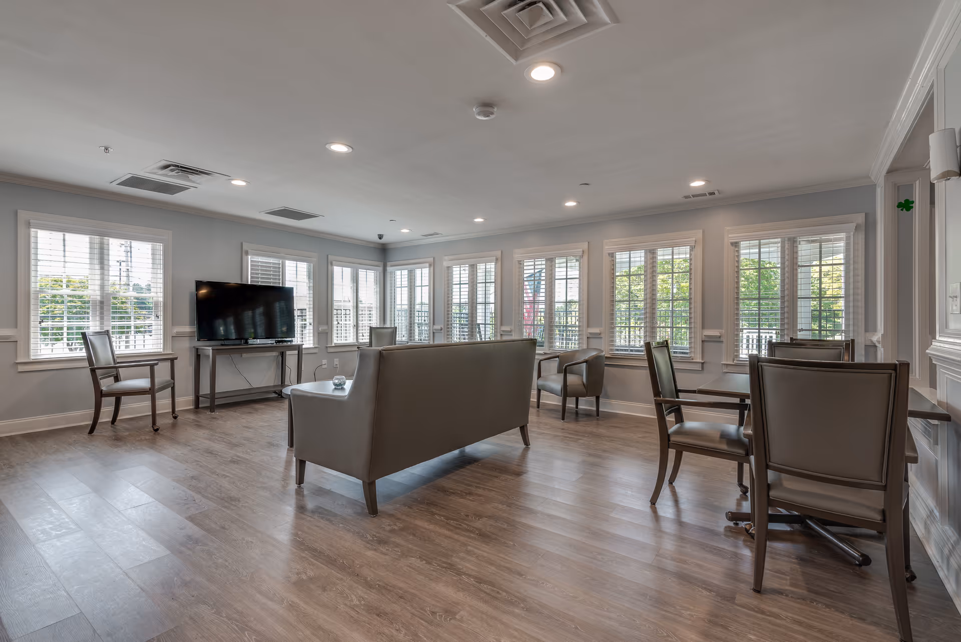 A spacious senior living common area with multiple windows letting in natural light. The room features a flat-screen TV on a stand, several chairs, a sofa, and tables arranged for seating. The floor is wood, and the walls are painted light gray with white trim.