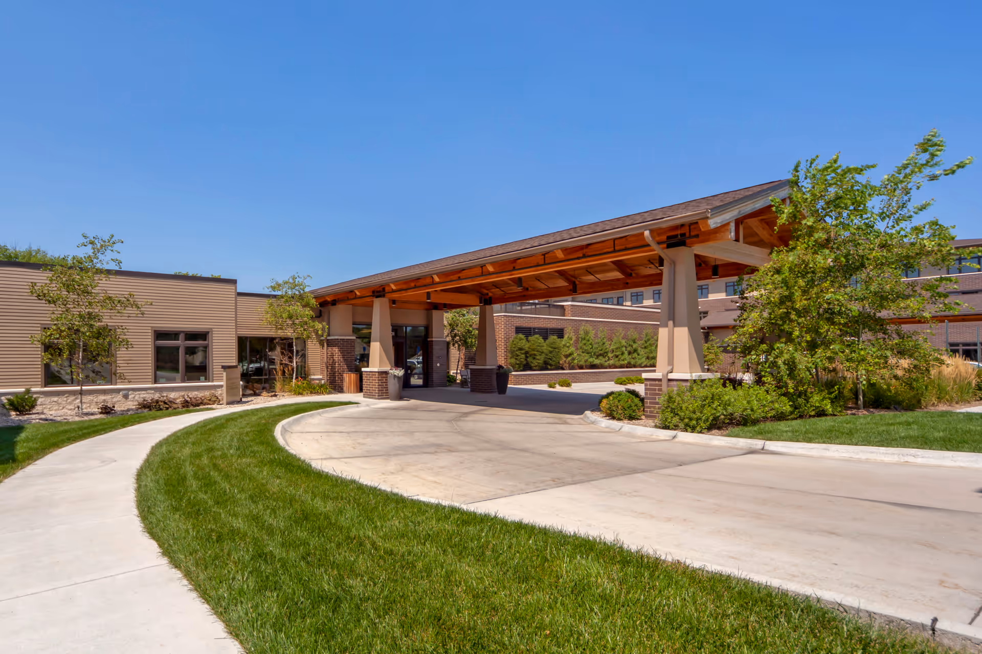 Entrance of The Knolls Senior Living facility showing a covered driveway with a wooden roof supported by pillars, surrounded by well-maintained green lawns, trees, and shrubs under a clear blue sky.