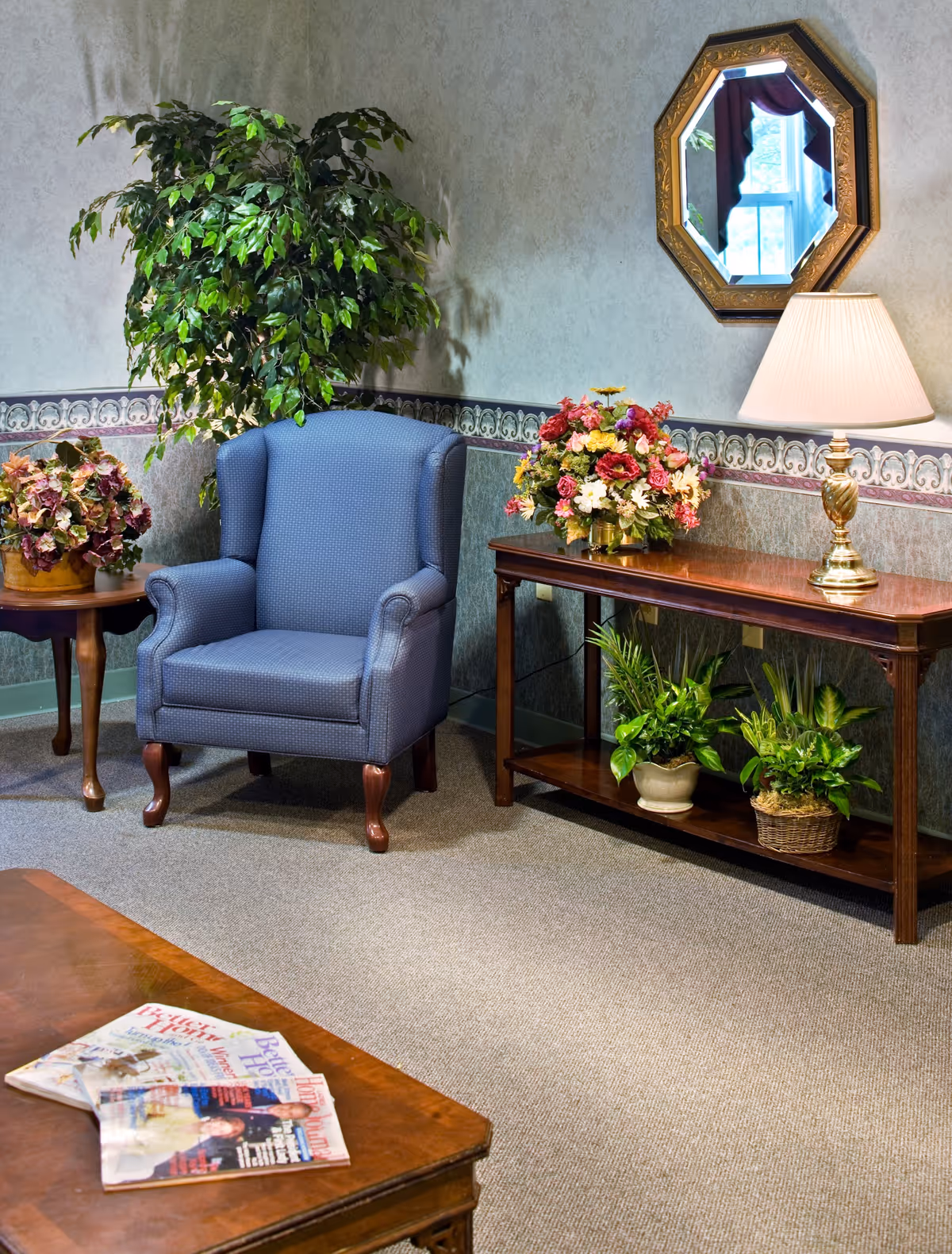 A cozy living room corner with a blue upholstered armchair, a wooden side table with a potted plant, a wooden console table with a floral arrangement and two potted plants underneath, a table lamp, and an octagonal mirror on the wall reflecting a window with curtains.