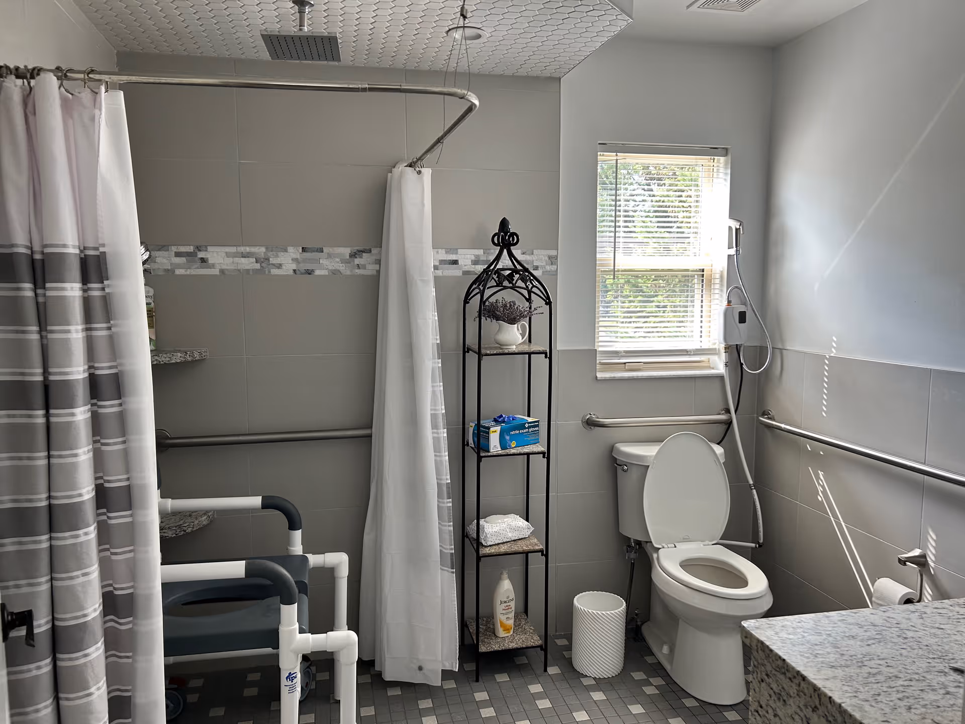 A clean and accessible bathroom with a toilet, a shower area with grab bars and a shower chair, a small shelving unit holding toiletries, a window with blinds, and a granite countertop. The walls are tiled and there are safety grab bars installed around the toilet and shower area.