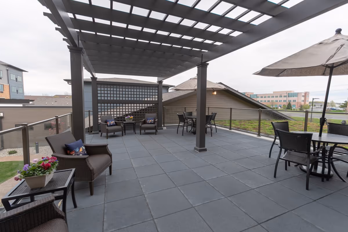 Outdoor patio area with a pergola overhead, featuring several wicker chairs with cushions, small tables with flower pots, and a round table with chairs under a large umbrella. The patio is surrounded by glass railing and overlooks nearby buildings and greenery.