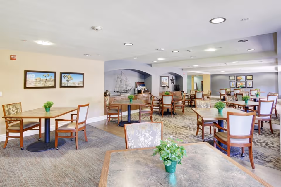 A spacious dining room with multiple wooden tables and chairs arranged neatly. Each table has a small green plant centerpiece. The walls are decorated with framed pictures and artwork, and the room is well-lit with ceiling lights.