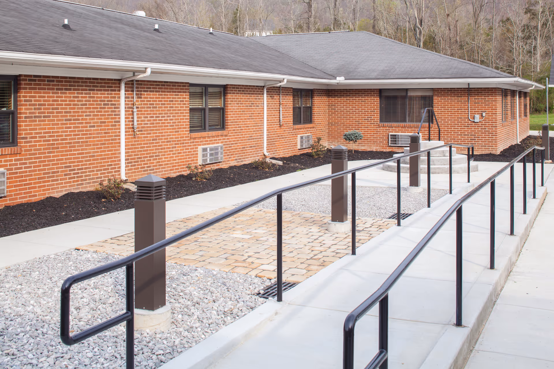 Wheelchair-accessible ramp with railings leading to the entrance of a single-story brick building with gravel and paver landscaping.