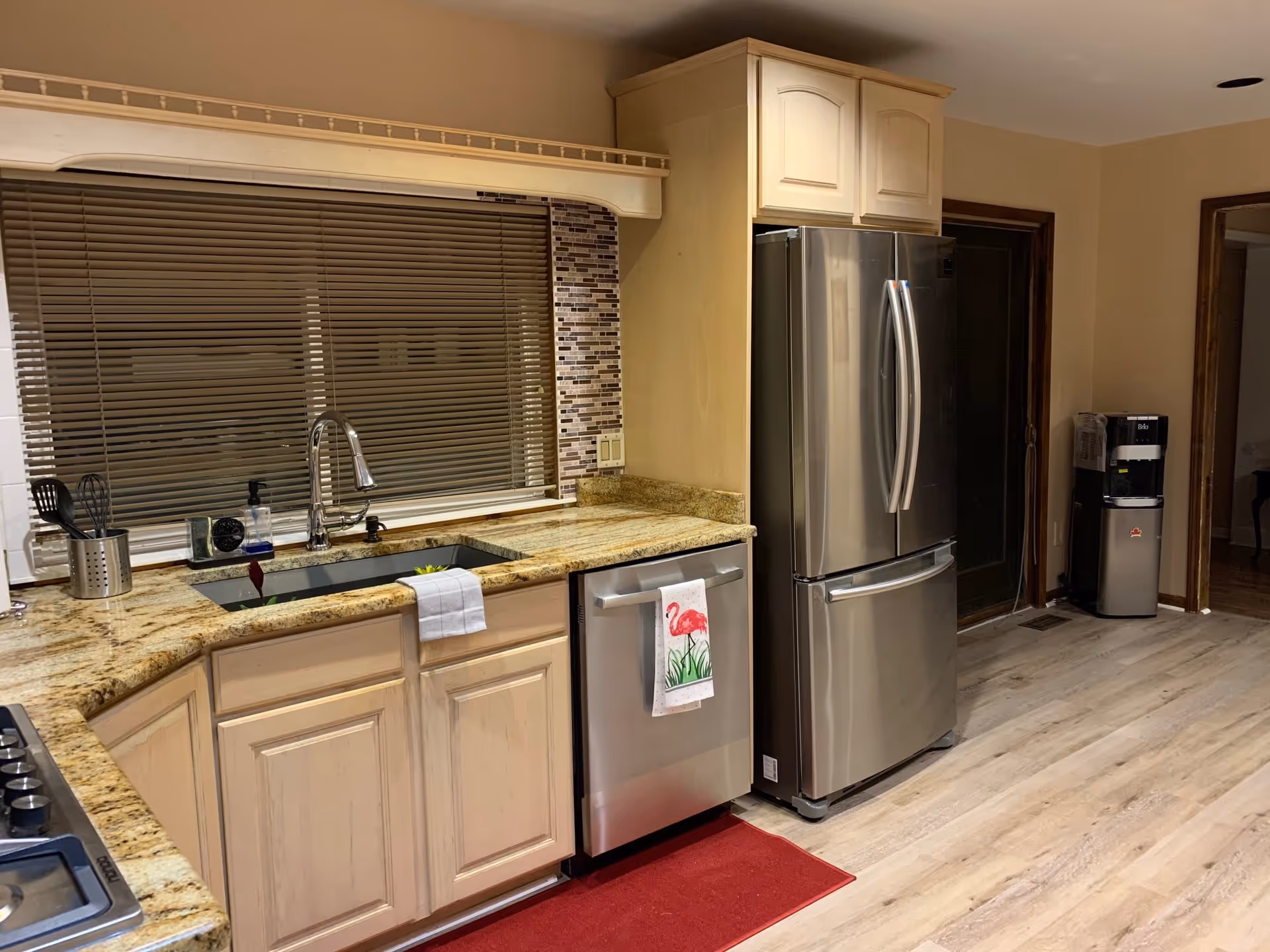 A modern kitchen with light wood cabinets, granite countertops, a stainless steel refrigerator, dishwasher, and a sink under a window with closed blinds. There is a red floor mat in front of the sink and dishwasher, and a water dispenser is visible in the background near a doorway.