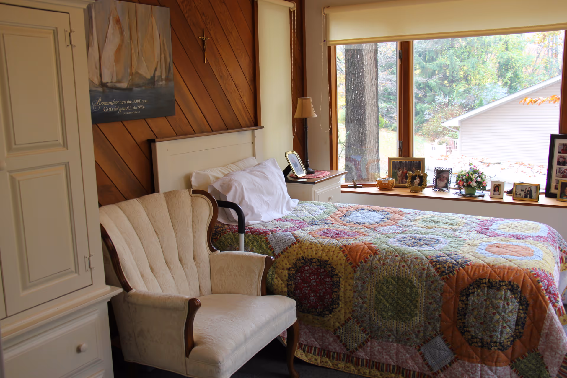 Sunlit bedroom with a quilt-covered bed, upholstered armchair, bedside table and large window displaying framed photos and an outdoor view.