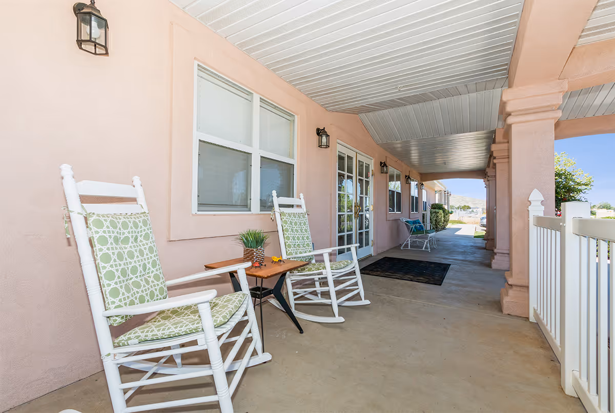 Covered outdoor patio area with two white rocking chairs featuring green patterned cushions, a small wooden table with a potted plant, and a white railing. The patio has a beige stucco wall with windows and a glass door, and a concrete floor extending into the distance with additional seating visible.