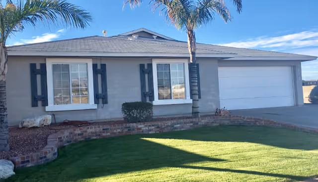 Front exterior of a single-story home with two shuttered windows, an attached garage, palm trees, and a manicured lawn.