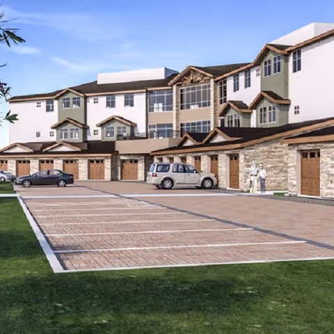 Exterior view of a multi-story senior living facility with attached garages on the ground level, several parked cars, and two people walking near the building. The building features a combination of stone and siding with multiple windows and a clear blue sky above.