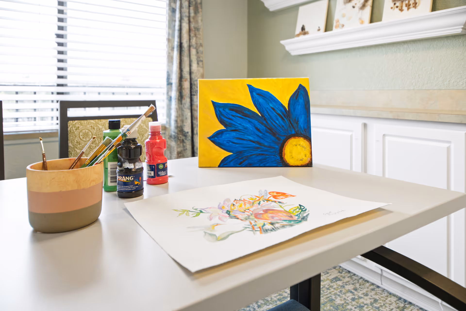 A table with painting supplies including paintbrushes in a cup, bottles of paint, and two paintings, one of a blue flower on a yellow background and another floral artwork on paper, set in a well-lit room with a window and white cabinets in the background.
