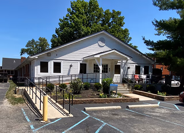 Front exterior of a single-story white care facility with a covered entrance, wheelchair ramp, steps, and parking in front.