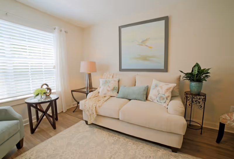 A cozy living room with a beige sofa adorned with floral and solid pillows, a soft throw blanket, a side table with a lamp, a round wooden table with a small flower arrangement, a green armchair, a potted plant on a decorative stand, and a framed painting of a bird in flight on the wall.