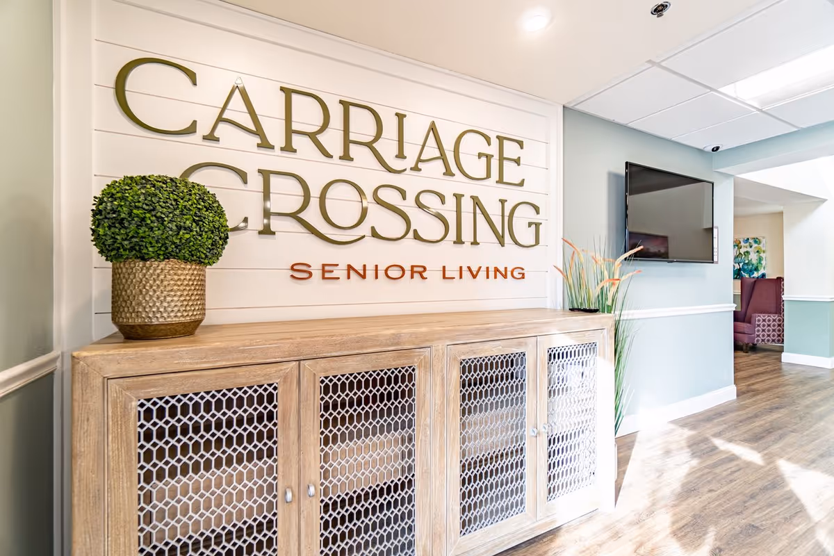 Interior view of a senior living facility reception or common area with a wooden cabinet featuring mesh doors, a potted plant on top, a wall sign that reads 'Carriage Crossing Senior Living', a mounted flat-screen TV, and a hallway leading to a seating area with purple chairs and artwork on the wall.