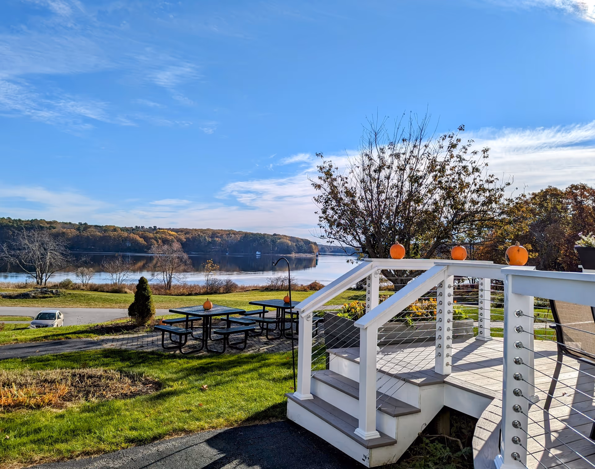 Outdoor view of a lakeside area with a clear blue sky, picnic tables with pumpkins on them, a white railing with pumpkins on top, green grass, and trees with autumn foliage.