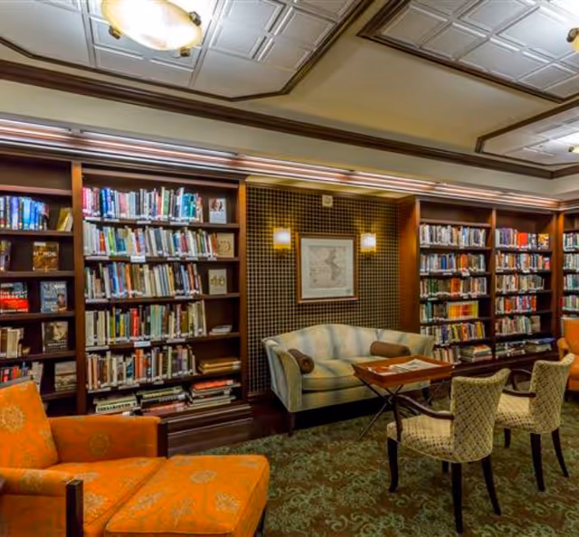 A cozy library room with wooden bookshelves filled with books along the walls. There is a light blue striped sofa with two brown cylindrical pillows, a wooden coffee table, two patterned chairs, and an orange armchair with a matching ottoman. The ceiling has decorative panels and a light fixture, and the walls have a patterned wallpaper with two wall sconces and a framed map.