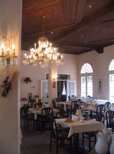 Interior view of a dining room with multiple tables covered in white tablecloths and set with folded napkins. The room features dark wooden chairs, a decorative wooden ceiling, large arched windows allowing natural light, and elegant chandeliers hanging from the ceiling.