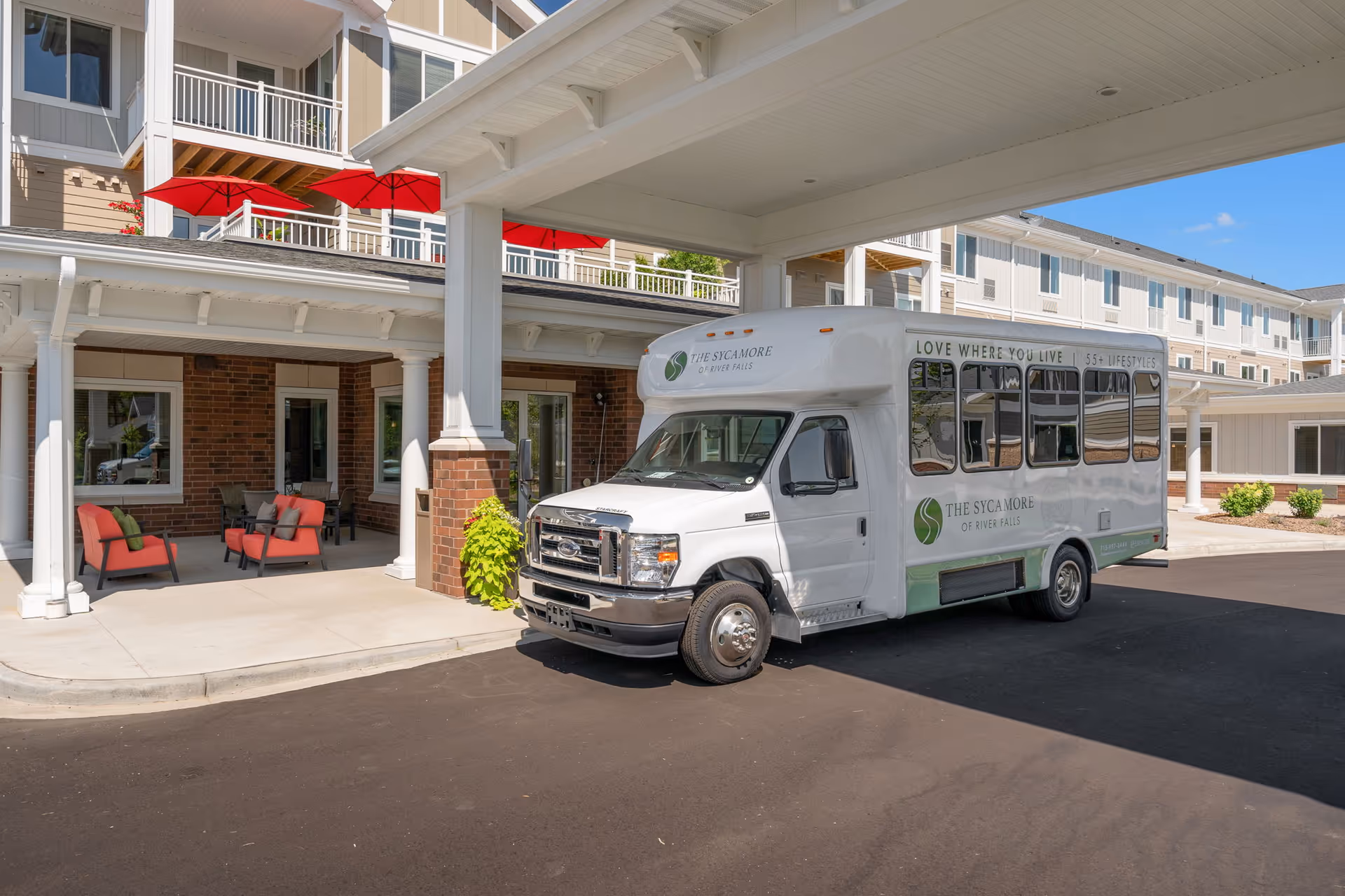 A white shuttle bus parked under the covered entrance of The Sycamore of River Falls senior living building, with outdoor seating and red umbrellas on balconies.