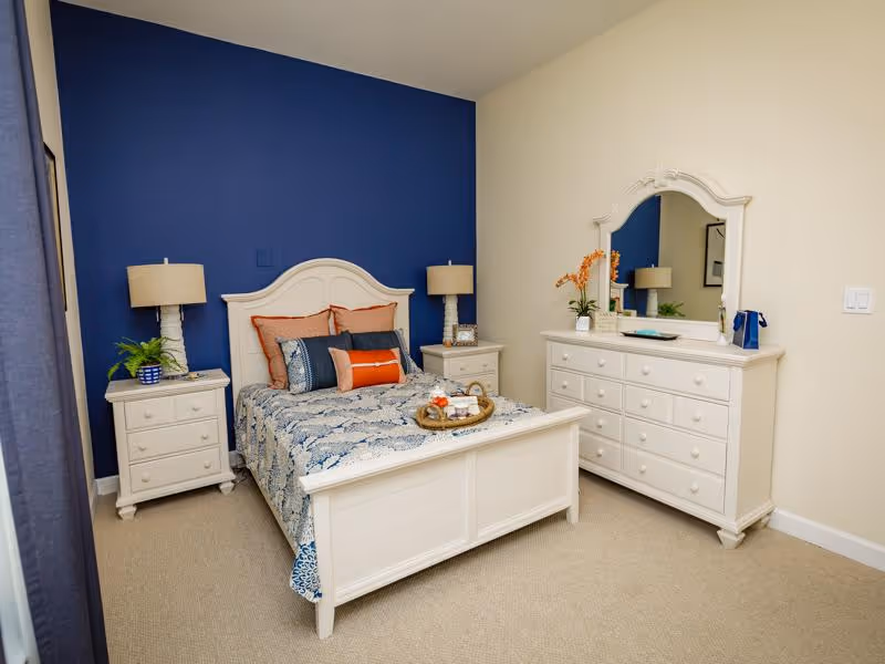 A neatly arranged bedroom with a white wooden bed frame and matching furniture including two nightstands and a dresser with a mirror. The bed is made with a blue and white patterned bedspread and several decorative pillows in shades of blue and orange. Two table lamps with beige shades sit on the nightstands. The wall behind the bed is painted dark blue, while the other walls are light beige. There are decorative plants and items on the nightstands and dresser.
