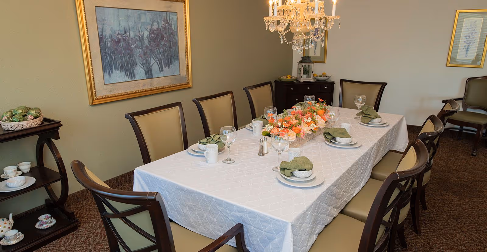 A formal dining room with a rectangular table covered with a white tablecloth, set with plates, bowls, green napkins, mugs, and wine glasses. A floral centerpiece decorates the middle of the table. The room has beige walls adorned with framed artwork, a chandelier hanging above the table, and a small side table with tea sets and a basket of artichokes.