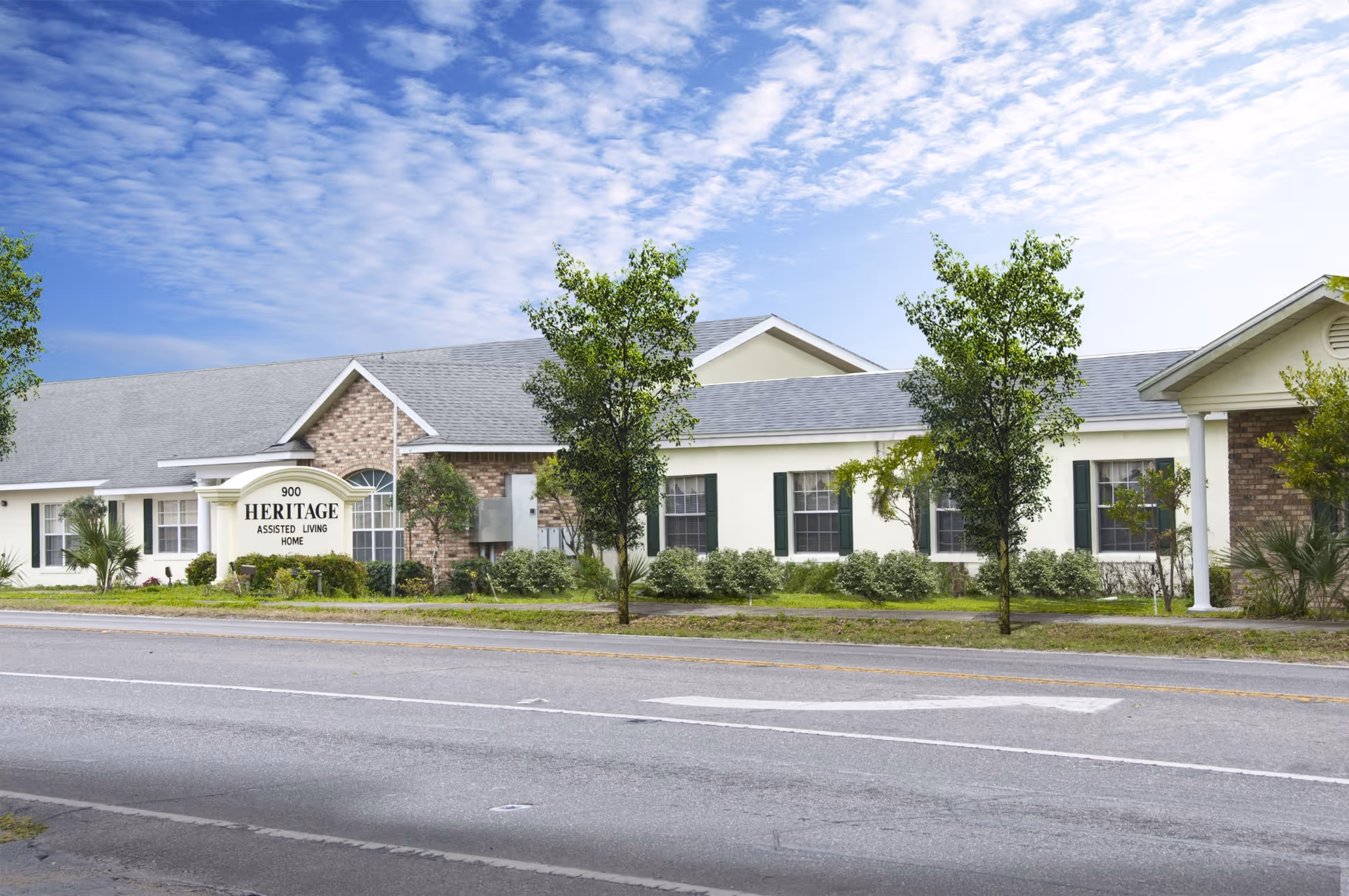 Front exterior of a single-story assisted living building with trees, landscaping, and a sign reading 'Heritage Assisted Living Home'.