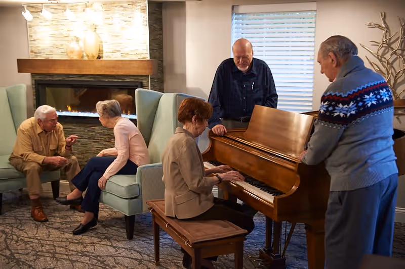 A group of five elderly people in a cozy living room setting. One woman is seated at a wooden piano playing, while two men stand nearby watching her. Another man and woman are seated on armchairs near a lit fireplace, engaged in conversation. The room has a warm and inviting atmosphere with soft lighting and a window with blinds.