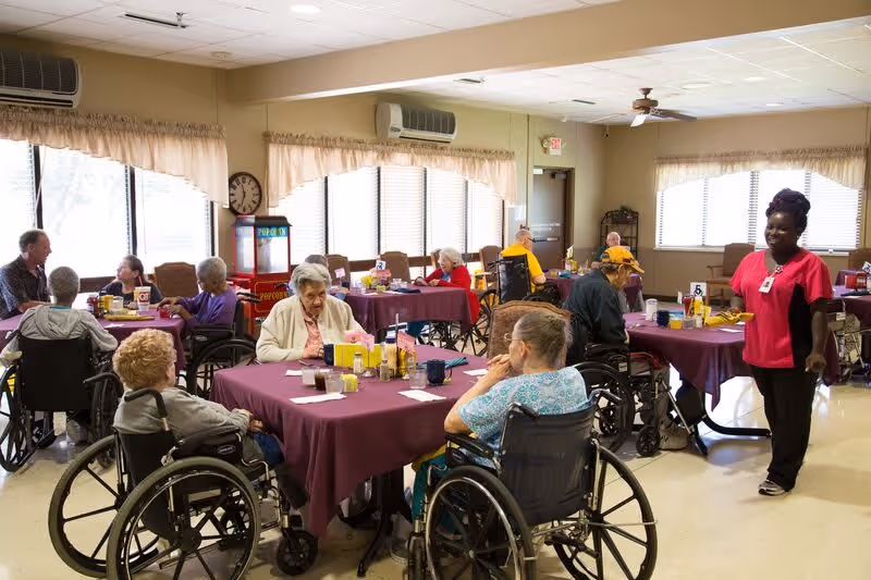 A dining room in a long term care facility with elderly residents sitting around tables covered with purple tablecloths. Most residents are in wheelchairs, eating or conversing. A staff member in a pink uniform is standing and smiling. The room has large windows with beige curtains, ceiling fans, and a popcorn machine in the corner.