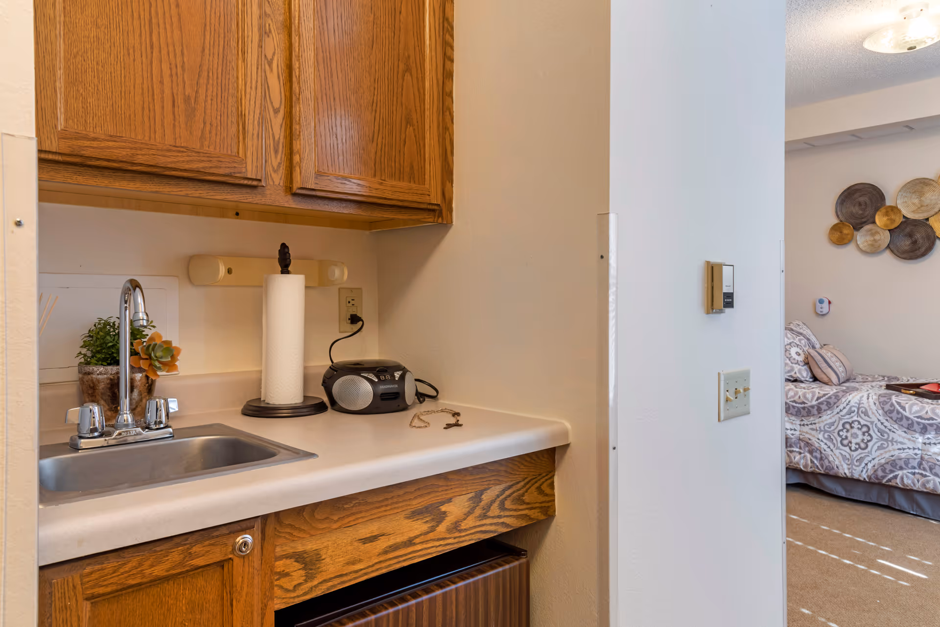 A small kitchenette area with a stainless steel sink, wooden cabinets above and below the countertop, a paper towel holder, a small radio, and a potted plant. To the right, a partial view of a bedroom with a bed covered in patterned bedding and decorative wall hangings is visible.