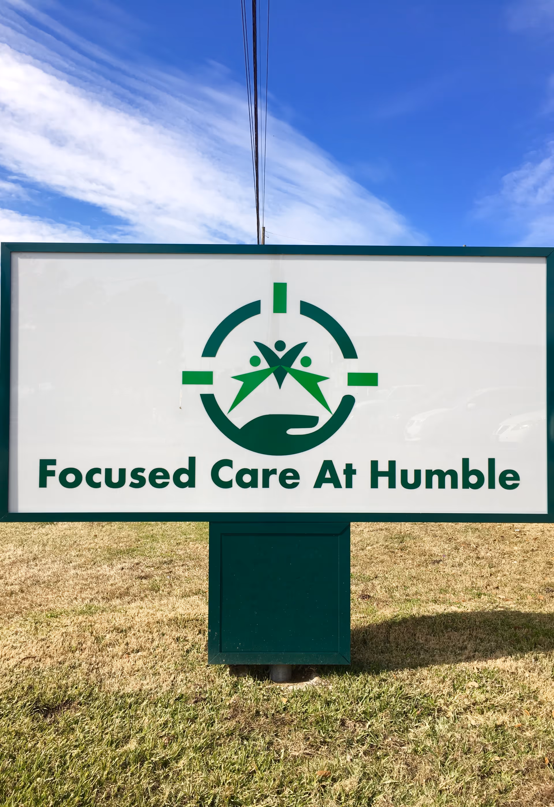 Outdoor sign for Focused Care At Humble with a green and white logo featuring three stylized human figures above an open hand, set against a blue sky with some clouds.