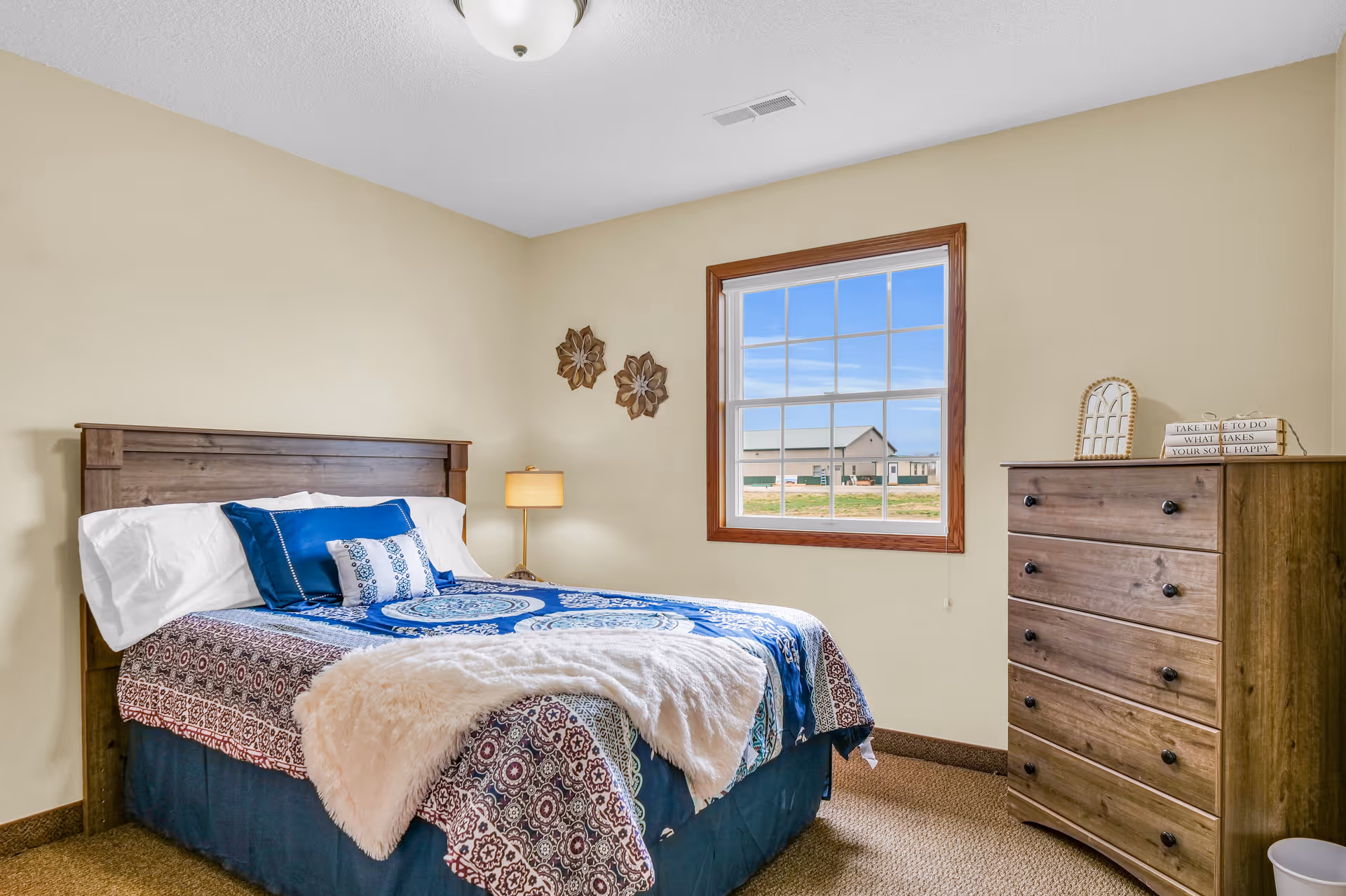 A cozy bedroom with a wooden bed frame and a bed made with blue and white patterned bedding, including several pillows. A soft white throw blanket is draped over the foot of the bed. There is a wooden dresser with five drawers on the right side of the room, topped with decorative items and a small sign. A window with wooden trim shows an outdoor view of buildings and a clear sky. The walls are painted beige, and a ceiling light fixture is visible.