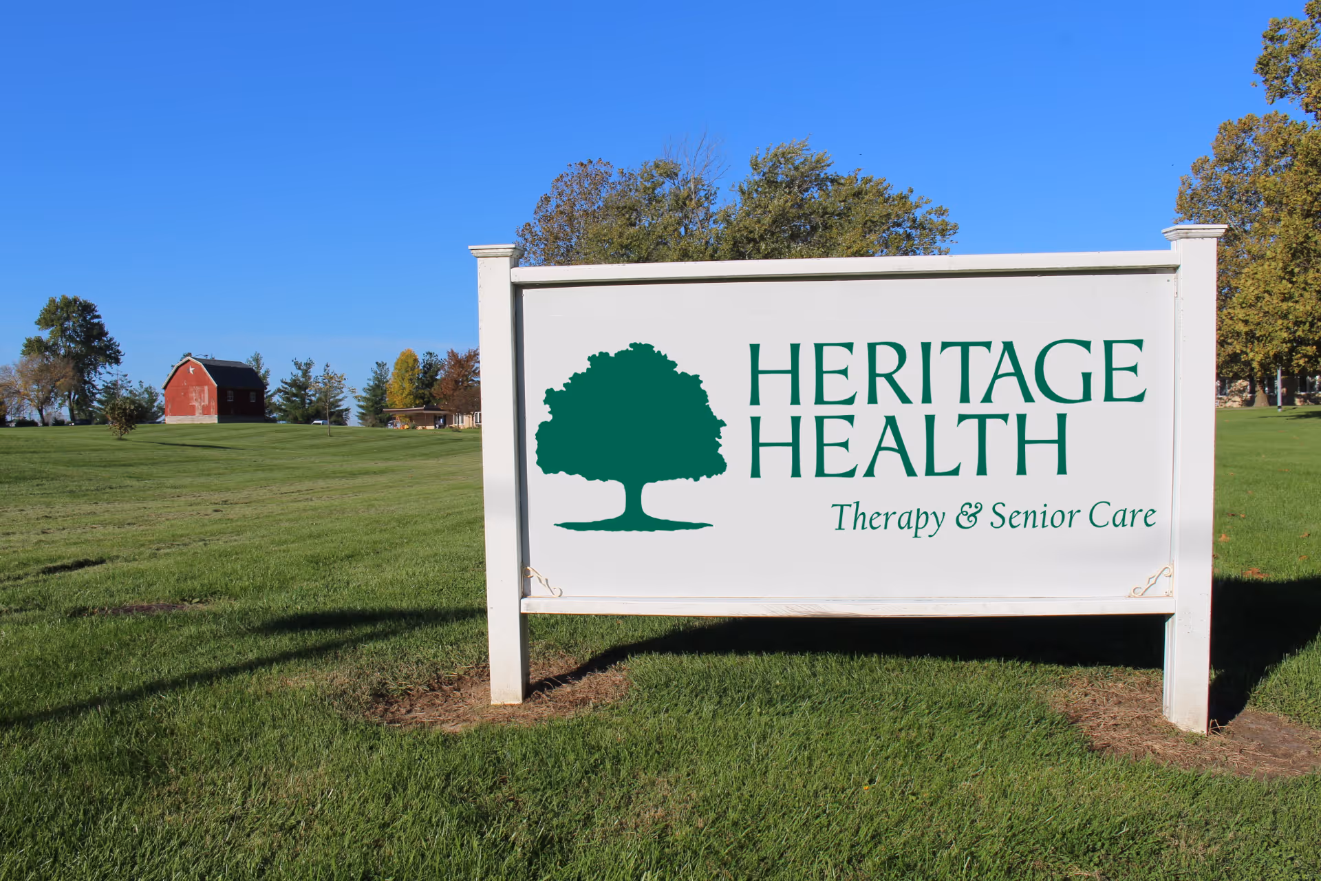 A large white sign with a green tree logo and the text 'HERITAGE HEALTH Therapy & Senior Care' stands on a grassy lawn with trees and a red barn in the background under a clear blue sky.