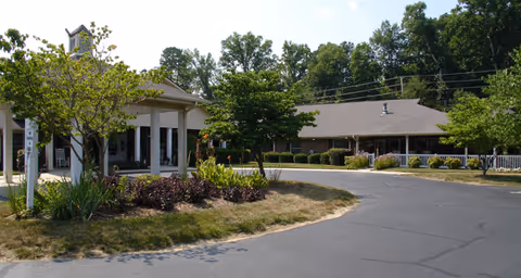 Front driveway and entrance of a single-story senior living building with a covered porte-cochere, landscaped beds, and surrounding trees.