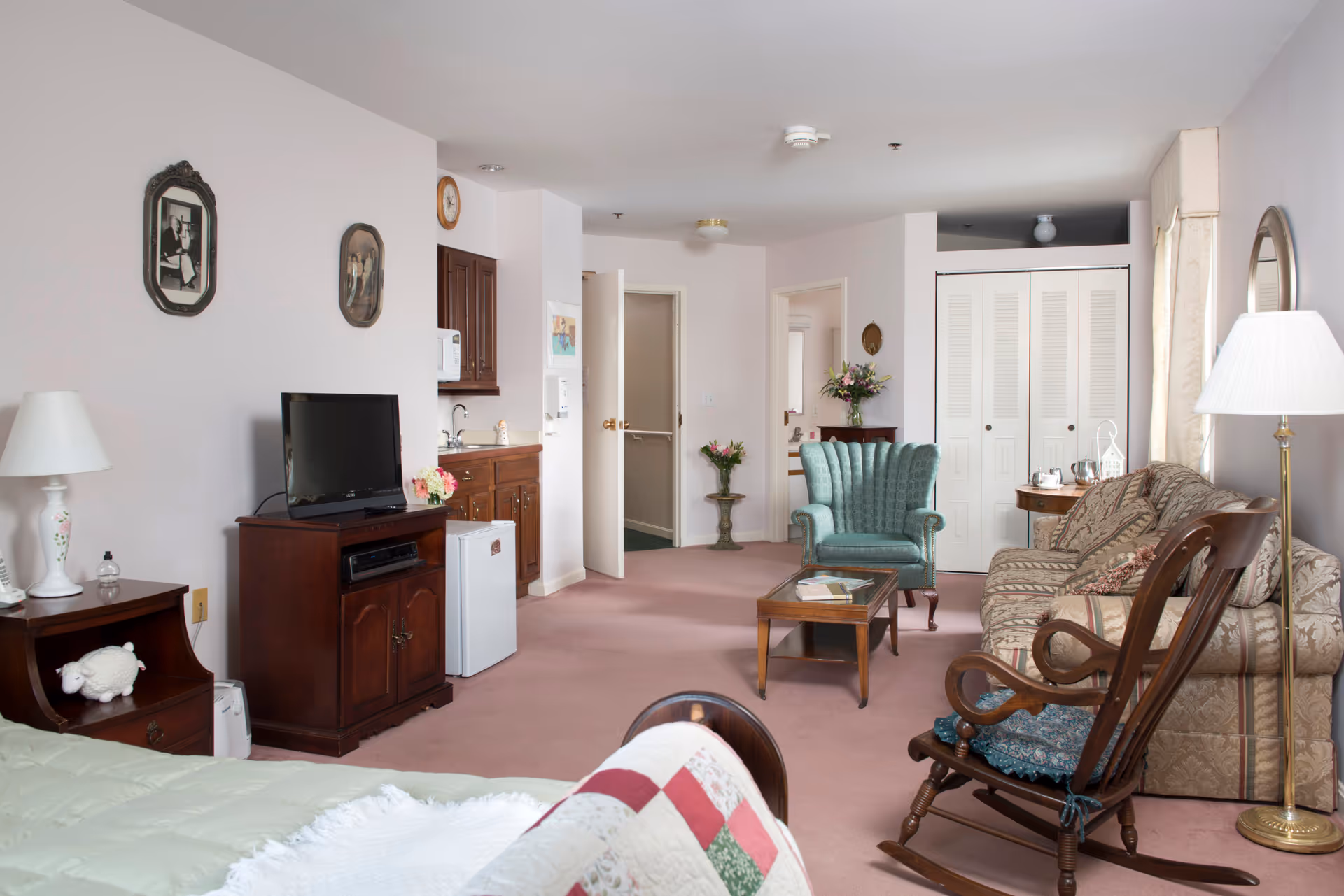A cozy senior living room with a patterned sofa, a green upholstered armchair, a wooden rocking chair, a wooden coffee table, and a TV on a wooden stand. The room has light pink carpet and walls, with framed pictures on the wall, a small kitchenette area with a microwave and mini fridge, and a bed partially visible in the foreground. There are lamps and floral decorations adding to the homey atmosphere.