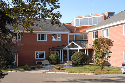 Exterior view of a two-story building with red siding and white trim, featuring multiple windows and a main entrance with a small covered porch. There are bushes, trees, and benches near the entrance, and a paved driveway in front.